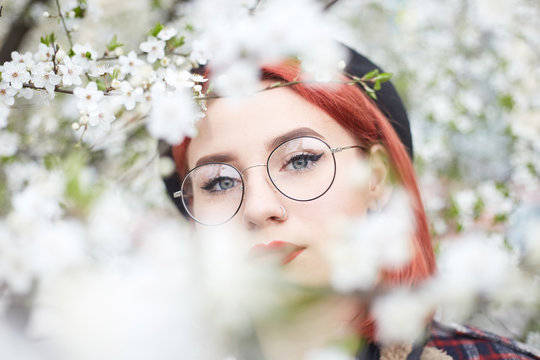 Outdoors fashion photo of young lady in round glasses and beret in the garden of cherry blossoms