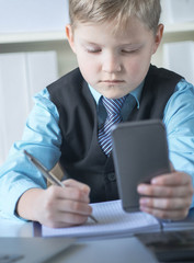 Serious little boy looking at the phone at father's table in office. Boy in business suit playing boss.
