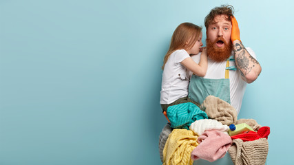 Overwhelmed surprised bearded ginger man keeps jaw dropped, keeps hand on head, carries crying daughter, feels puzzled, has to wash laundry, stands against blue studio wall with blank space.