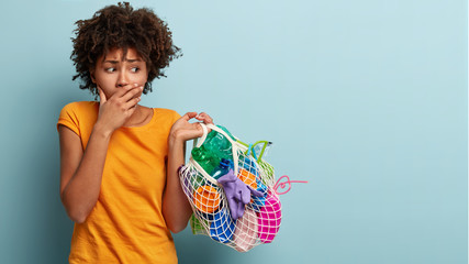 Nervous black woman with curly hair, covers mouth, looks worried at bag of plastic trash, shows...