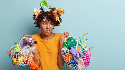 Horizontal shot of discontent dark skinned Afro American female frowns face as picks up garbage,...