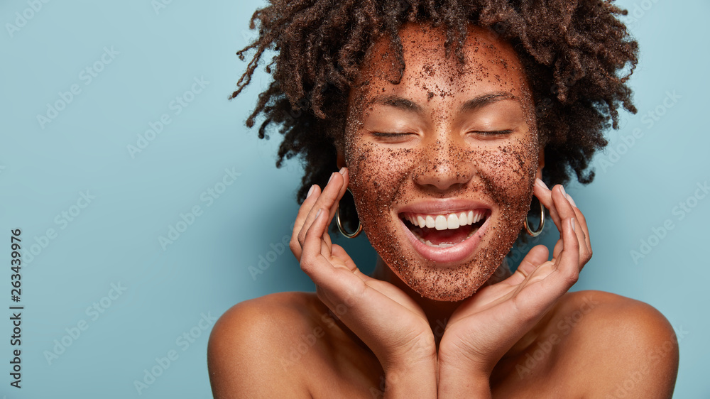 Poster Cheerful dark skinned lady applies coffee scrub on face, pampers skin, closes eyes from pleasure, smiles positively has bare shoulders, stands against blue background, free space aside. Beauty concept