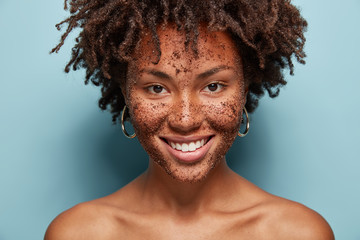 Beautiful mixed race female has skin scrub on face, smiles gently and looks directly at camera, makes cosmetic masks from coffee, has curly hairstyle, bare shoulders, isolated over blue background