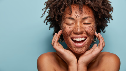 Cheerful dark skinned lady applies coffee scrub on face, pampers skin, closes eyes from pleasure, smiles positively has bare shoulders, stands against blue background, free space aside. Beauty concept - obrazy, fototapety, plakaty
