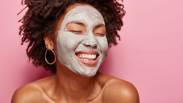 Close up portrait of happy delighted woman applies clay mask, closes eyes and smiles delightfully, imagines something pleasant, stands over pink background empty space on right side. Skin care concept
