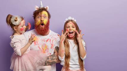 Horizontal shot of shocked dad has yellow face painted with watercolors, two children have fun with father, joyful expressions, isolated over purple background with free space for promotion.