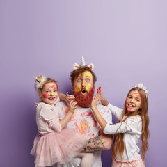 Vertical shot of astonished red haired man wears unicorn horn, plays with two small girls, have fun...