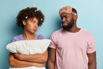 Upset displeased dark skinned husband and wife look with sad gloomy facial expressions at each other, have bad mood after sleeping for short time, wears sleepmask, isolated over blue background.