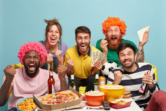 Group of mixed race best friends watch football match with excitement, shout for favourite team, make sport betting on money, clench fists, eat pizza, popcorn, drink beer, celebrate goal, cheer up
