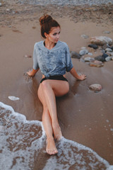 Young slim tanned woman in black swimsuit and grey t-shirt sitting on sand near the sea in the evening after sunset. Girl enjoys the sea waves.