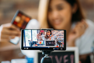 Happy young attractive Asian ethnic model filming vlog on live stream on mobile phone on tripod and showing eyeshadows at table with cosmetics