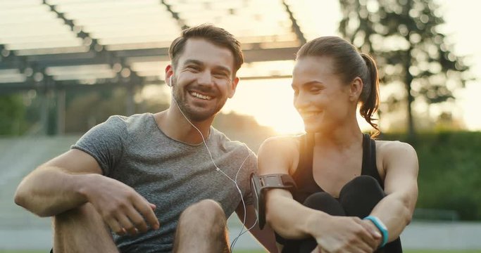 Happy Caucasian young couple in love sitting and resting after workout at the stadium, laughing and listening to the music. Portrait.