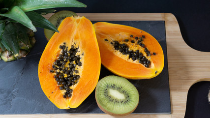 The papaya is cut into two halves, the stand is a stone board with a wooden frame, near the Papay there is a cut off top of the pineapple, the background is black