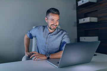 Smiling hardworking Caucasian businessman looking at laptop while sitting in office late at night.