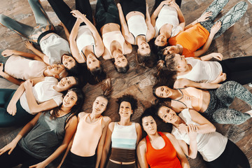 Top view of sporty group of women in sportswear lying down on gym floor in circle. Fit is not...