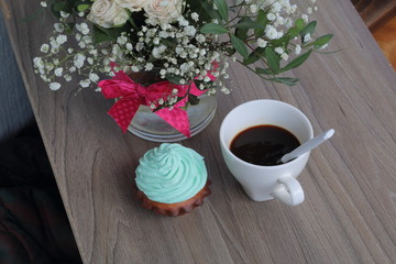 Cream cake and cup of coffee. A coffee spoon is dropped into the coffee cup. Nearby is a bouquet of flowers in a glass vase.