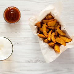 Fried potato wedges in paper box, barbecue sauce and glass of cold beer on a white wooden surface. Flat lay. Overhead, top view, from above.