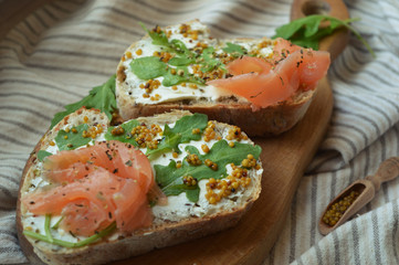 Homemade salmon sandwiches with cream cheese, mustard seeds, olive oil, arugula on toasted rye bread, served on the wooden board. Healthy trendy food. Delicious breakfast.