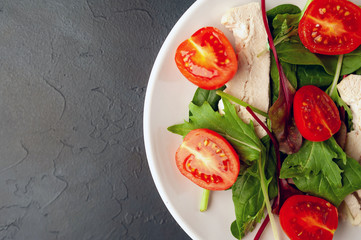 Mix fresh leaves of arugula, lettuce, spinach, tomato and chicken fillet for salad, on a white plate on a stone background with copy space for your text