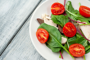 Mix fresh leaves of arugula, lettuce, spinach, tomato and chicken fillet for salad, on a white plate on a wood background