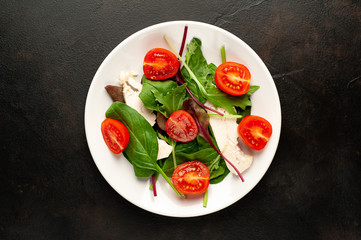 Mix fresh leaves of arugula, lettuce, spinach, tomato and chicken fillet for salad, on a white plate against a stone