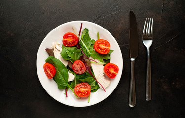 Mix fresh leaves of arugula, lettuce, spinach, tomato and chicken fillet for salad, on a white plate against a stone