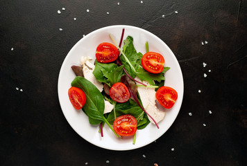 Mix fresh leaves of arugula, lettuce, spinach, tomato and chicken fillet for salad, on a white plate against a stone