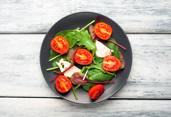 Mix fresh leaves of arugula, lettuce, spinach, tomato and chicken fillet for salad, on a dark plate on a wood background