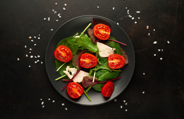 Mix fresh leaves of arugula, lettuce, spinach, tomatoes and chicken fillet for salad, on a dark plate against a background of concrete