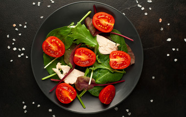 Mix fresh leaves of arugula, lettuce, spinach, tomatoes and chicken fillet for salad, on a dark plate against a background of concrete