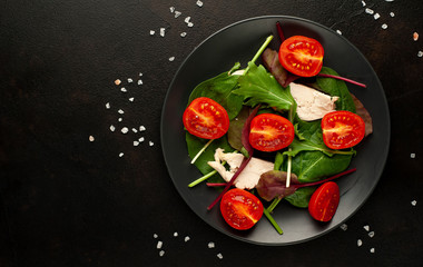 Mix fresh leaves of arugula, lettuce, spinach, tomatoes and chicken fillet for salad, on a dark plate against a background of concrete
