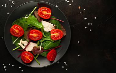 Mix fresh leaves of arugula, lettuce, spinach, tomatoes and chicken fillet for salad, on a dark plate against a background of concrete