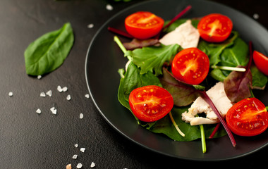 Mix fresh leaves of arugula, lettuce, spinach, tomatoes and chicken fillet for salad, on a dark plate against a background of concrete