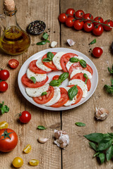 Delicious italian caprese salad with ripe tomatoes, fresh basil and mozzarella cheese on wooden rustic background. Italian caprese salad in white plate with sliced tomatoes.