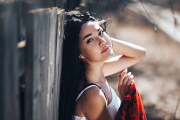 Portrait of a Beautiful black haired girl in a white vintage dress standing near wood fence.Young woman model posing in a russian national style . red shawl