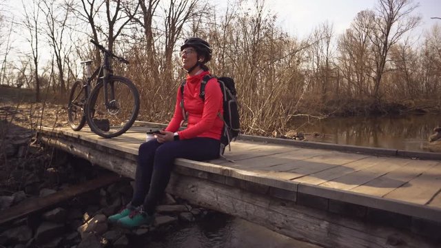 Happy young woman sitting wooden bridge over river, listening music in headphones with smartphone hands and dancing. Positive cyclist listens song in nature. Athlete happy near bike ears earphones