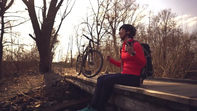 Happy young woman sitting wooden bridge over river, listening music in headphones with smartphone hands and dancing. Positive cyclist listens song in nature. Athlete happy near bike ears earphones