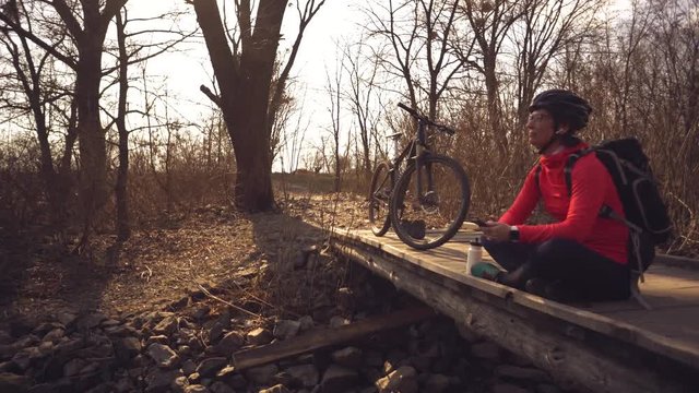 Happy young woman sitting wooden bridge over river, listening music in headphones with smartphone hands and dancing. Positive cyclist listens song in nature. Athlete happy near bike ears earphones