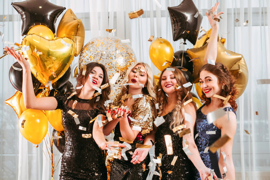 Girls party. Festive event. Ladies in cocktail dresses throwing confetti, posing over big window decorated with golden balloons.