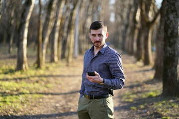 Portrait of a serious business bearded man with freckles with short hair in a plaid shirt, olive pants, holding a phone in his hand and looking right in the autumn forest among the trees in the sun.