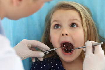Little baby girl sitting at dental chair with open mouth and feeling fear during oral check up while doctor. Visiting dentist office. Medicine concept