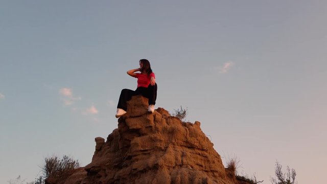Bellissima ragazza seduta su una sporgenza di terra fa oscillare il piede allegramente. Maglietta rosa, pantaloni neri e scarpe bianche. Vista dal basso.