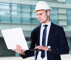 Smiling architector in suit and hat is exploring project in his laptop