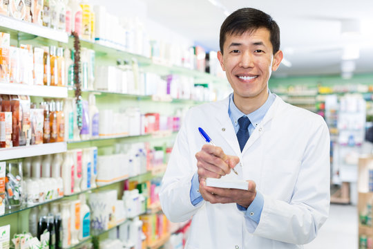 Chinese male looking medicine with notebook near shelves in pharmacy
