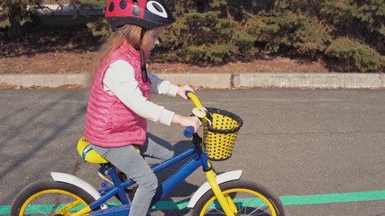 Little cute blond girl in protective helmet riding blue bike on an asphalt track with gaps and holes in old park. Sport concept, active happy kid playing and cycling first bicycle outdoors.