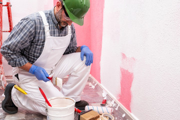 Caucasian house painter worker in white overalls, with helmet and goggles he prepares the white paint to paint the pink wall. Construction industry. Work safety.