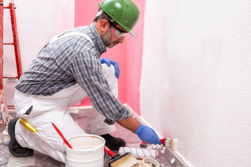 Caucasian house painter worker in white overalls, with helmet and goggles painting the pink wall with white paint using a small brush. Construction industry. Work safety.