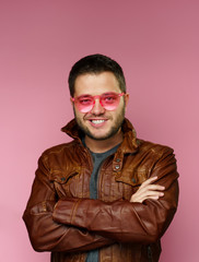 Obraz premium Photo of smiling brunette with pink glasses, leather jacket, with arms crossed on pink background.