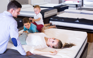 Young attractive woman with husband and sons lying on back on tested mattress in store