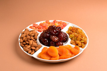 Mix of dried fruits and nuts on a white plate. Apricot, almond, raisin, dates fruit. On a brown background. Space for text or design.
